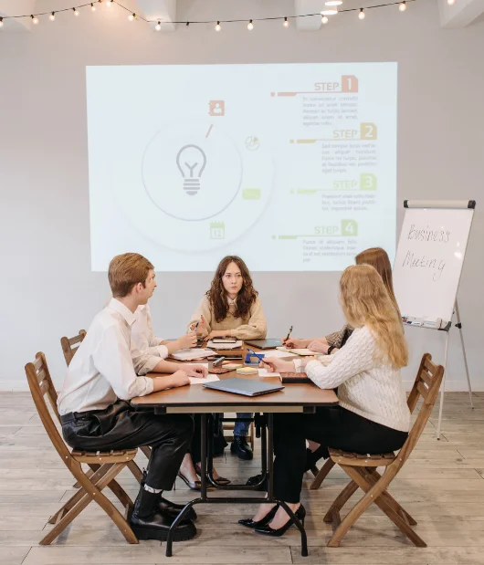 A group of professionals in a business meeting discussing a step-by-step strategy, with a presentation slide displaying a lightbulb and four steps.