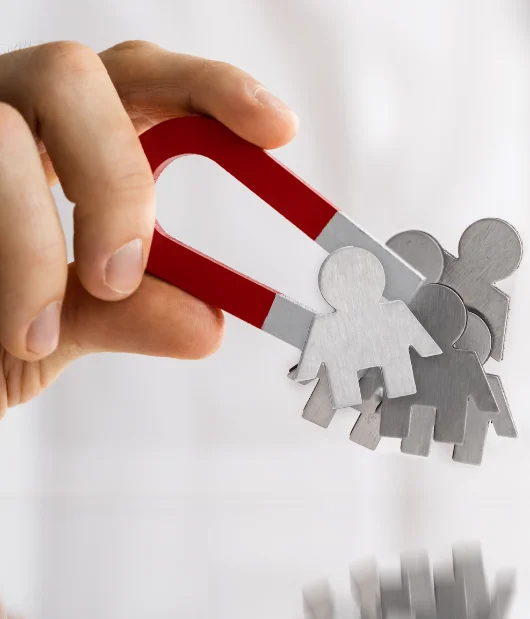 A close-up of a hand holding a red and silver magnet pulling metallic human figures, representing customer attraction and lead generation.